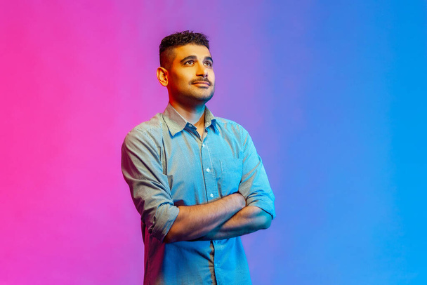 Portrait of calm handsome young man in shirt standing with crossed arms, looking away with dreamy facial expression. Indoor studio shot isolated on colorful neon light background. - Foto, immagini