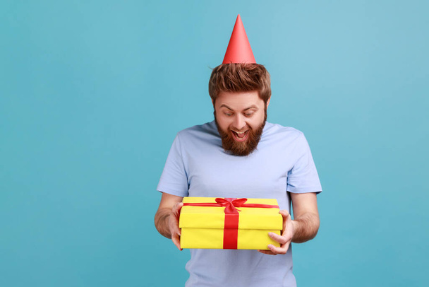 Portrait of excited surprised bearded man in party cone holding yellow wrapped gift box, looking at present with satisfied expression. Indoor studio shot isolated on blue background. - Zdjęcie, obraz