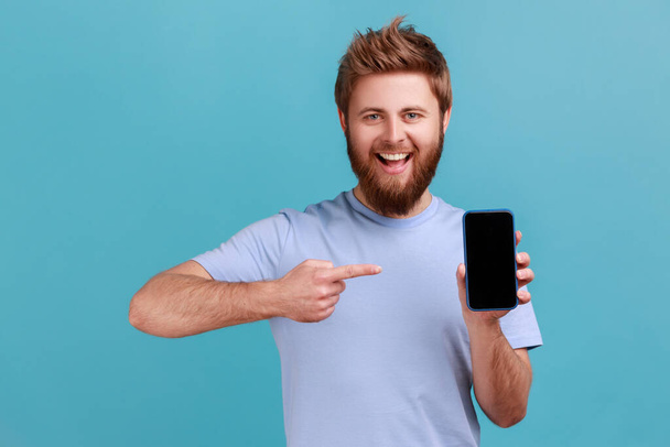 Portrait of smiling happy bearded man pointing finger at smartphone with black blank screen, copy space for advertisement or promotion. Indoor studio shot isolated on blue background. - 写真・画像