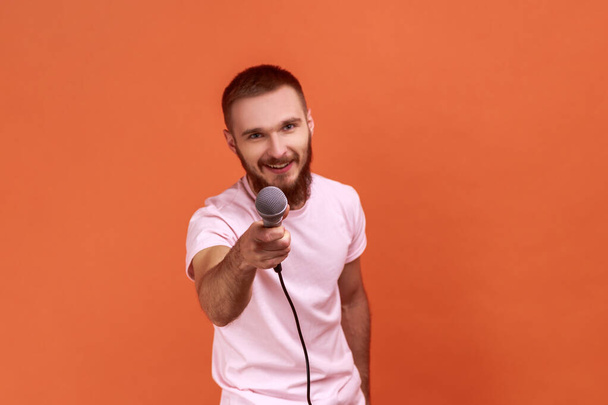Portrait of bearded man posing with microphone in hands, offers mic, journalist asking questions, looking at camera, wearing pink T-shirt. Indoor studio shot isolated on orange background. - Photo, image