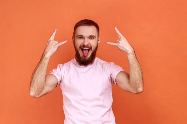 Portrait of bearded man showing rock and roll sign hand gesture, looking at camera with tongue out, excited expression, wearing pink T-shirt. Indoor studio shot isolated on orange background. - Zdjęcie, obraz