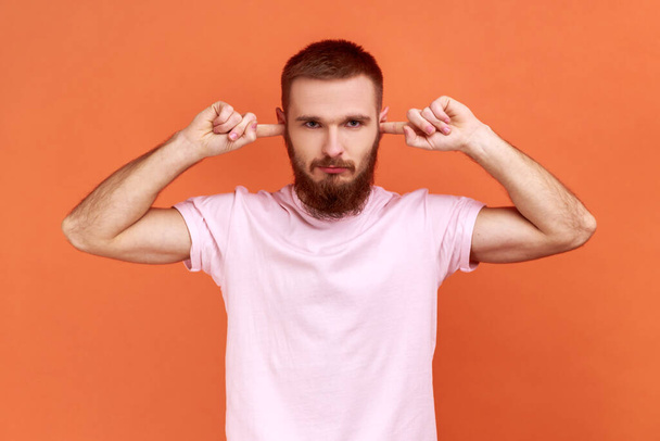 Portrait of bearded man closing ears with fingers seriously looking at camera, tired of irritated noise, wants silence, wearing pink T-shirt. Indoor studio shot isolated on orange background. - Zdjęcie, obraz