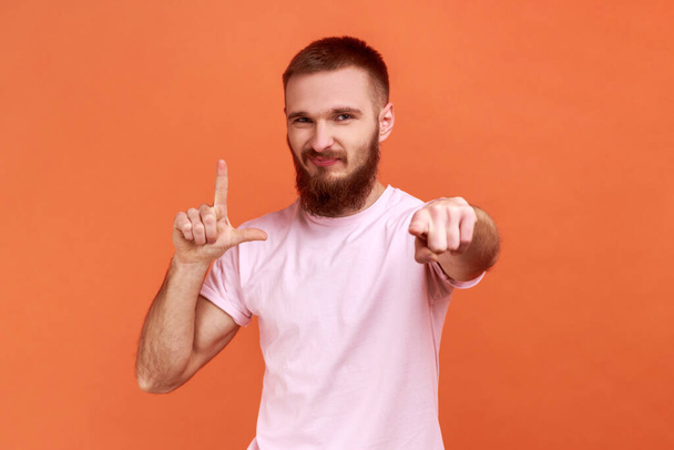 Hey, you failed. Portrait of bearded man showing loser gesture, pointing to camera, accusing for mistake, expressing disrespect, wearing pink T-shirt. Indoor studio shot isolated on orange background. - Foto, Imagen