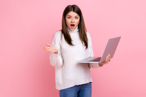 Portrait of attractive shocked girl standing with laptop in hands, looking at camera with open mouth, wearing white casual style sweater. Indoor studio shot isolated on pink background. - Photo, image