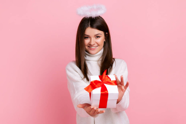 Charming brunette angelic woman with nimb over head, giving present box, congratulating with holiday, wearing white casual style sweater. Indoor studio shot isolated on pink background. - Photo, Image