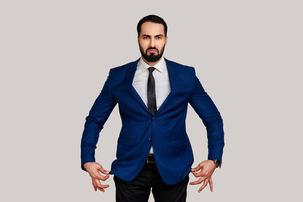 Portrait of sad unhappy bearded man showing empty pockets inside out, going bankrupt, no cash for living, wearing official style suit. Indoor studio shot isolated on gray background. - Фото, изображение