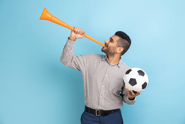 Delighted businessman celebrating victory of favourite football team, blowing horn and holding ball in hands, wearing striped shirt. Indoor studio shot isolated on blue background. - Valokuva, kuva