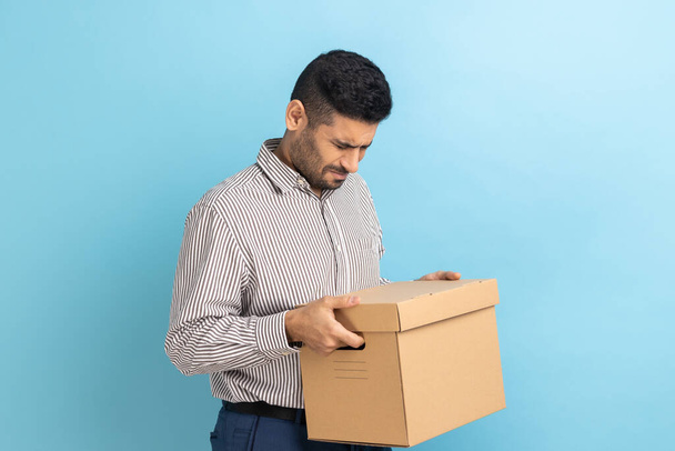 Portrait of positive businessman holding cardboard parcel, delivering orders to address, looking at carton package, wearing striped shirt. Indoor studio shot isolated on blue background. - Foto, immagini