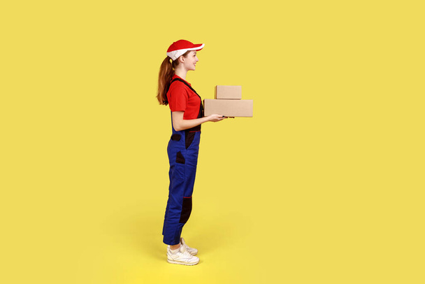 Side view portrait of courier woman standing with two parcels in hands, looking ahead, delivery service, wearing overalls and red cap. Indoor studio shot isolated on yellow background. - Fotó, kép