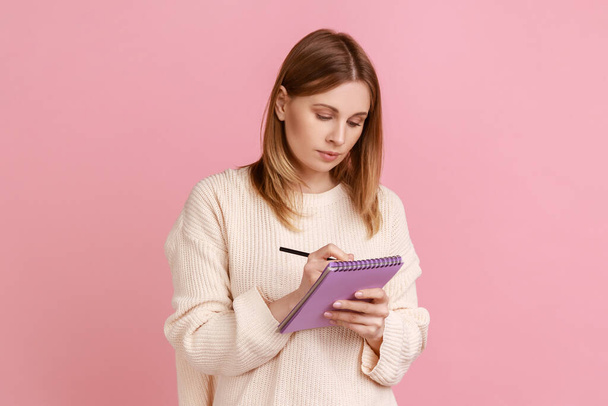 Portrait of serious concentrated blond woman writing in paper notebook, making to do list or list of purchases, wearing white sweater. Indoor studio shot isolated on pink background. - Фото, изображение