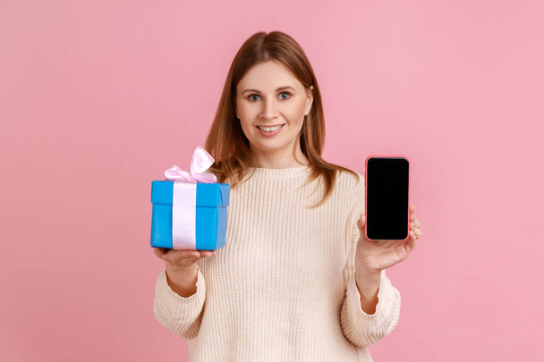 Smiling happy blond woman holding wrapped present box and smart phone with empty screen for advertisement and promotional text, wearing white sweater. Indoor studio shot isolated on pink background. - Photo, image