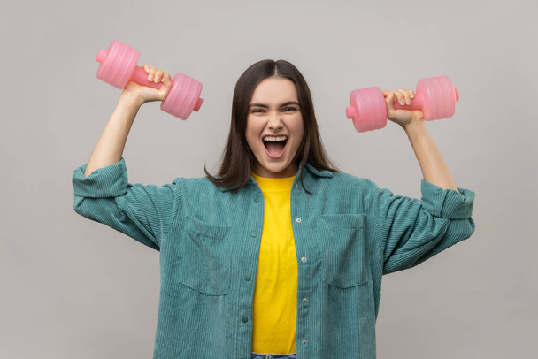 Woman with dark hair holding pink dumbbells in hands and looking at camera with excited expression, pumping up muscles, wearing casual style jacket. Indoor studio shot isolated on gray background. - Фото, изображение