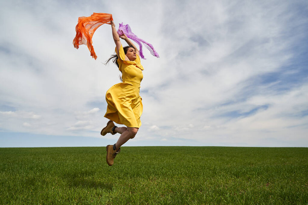 Happy young woman in yellow dress jumping outdoor on a green wheat field in a sunny day - Zdjęcie, obraz