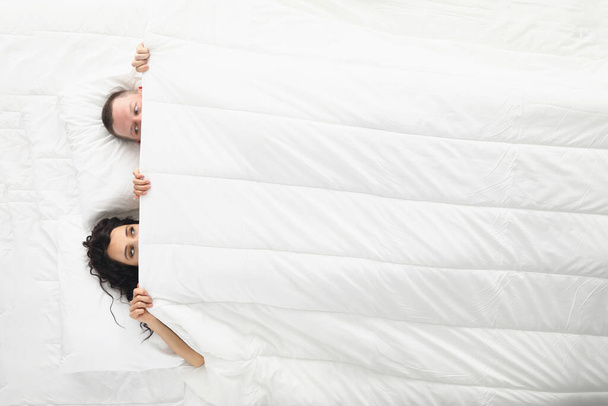 Woman and man peeking on each other under white blanket, awake couple in bed - Foto, Imagem