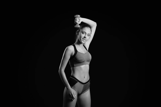 Черно-белый портрет красивой спортивной молодой женщины с гантелями на темном фоне - Фото, изображение