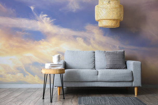 Зручний диван зі столом і стильна люстра біля стіни з надрукованим небом
 - Фото, зображення