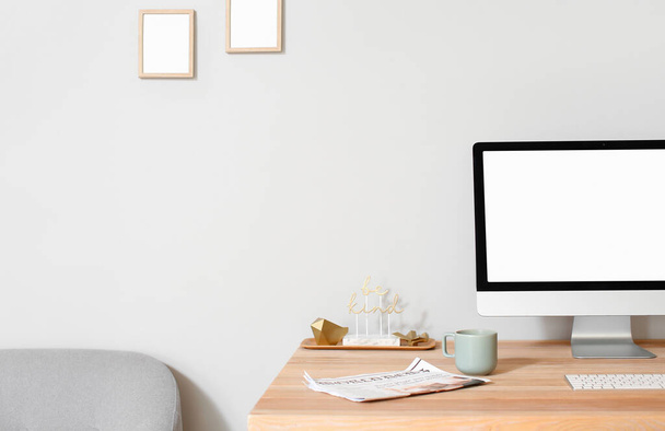 Ordinateur moderne, journal, tasse et décor avec texte BE KIND sur un bureau en bois près d'un mur lumineux - Photo, image