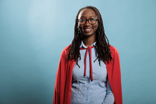Молодая очаровательная красивая улыбающаяся африканская супергероиня женщина в красном плаще на синем фоне. - Фото, изображение