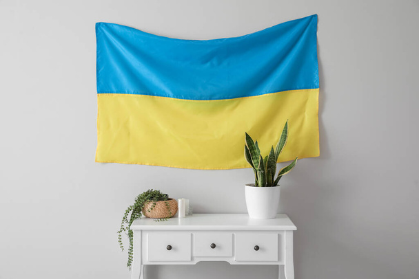 Table with houseplants, candles and Ukrainian flag hanging on light wall - Фото, зображення