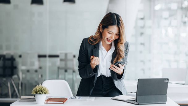 Woman gesturing and looking at mobilephone screen, businesswoman checking company monthly sales and pretending to be happy as sales meet planned targets according to policy. Sales management concept. - 写真・画像