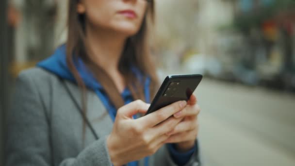 Γυναίκα με γυαλιά στέκεται σε στάση δημόσιων συγκοινωνιών και χρησιμοποιεί smartphone. Τραμ περνά στο παρασκήνιο. - Πλάνα, βίντεο