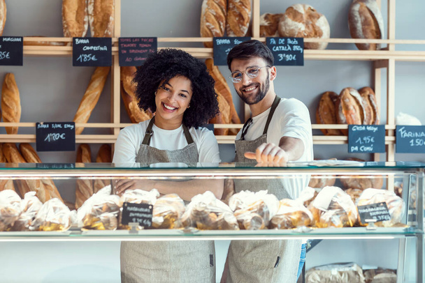 Portrait of two younger owners selling fresh pastry and loaves un bread section and smiling at pastry shop. - Foto, Imagem