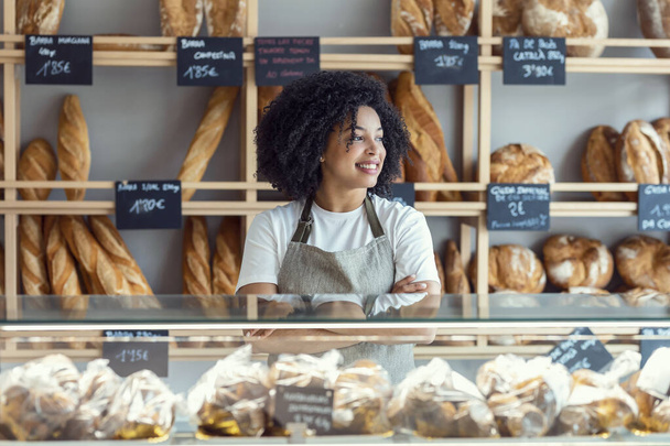 Portrait of beautiful afro younger owner woman selling fresh pastry and loaves un bread section and smiling at pastry shop. - 写真・画像