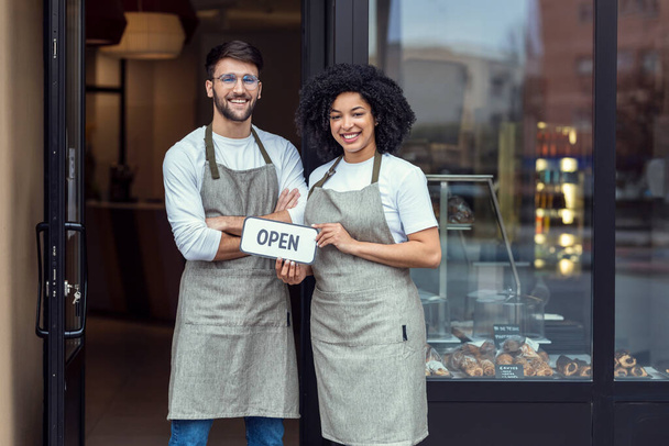 Portrait of two owners friends holding a open sign together while smiling to the camera in front of pastry shop. - Φωτογραφία, εικόνα