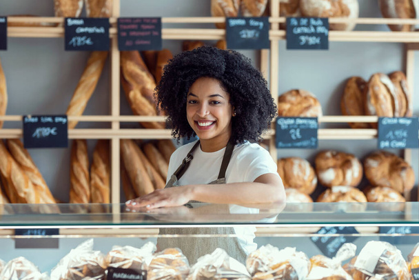 Portrait of beautiful afro younger owner woman selling fresh pastry and loaves un bread section and smiling at pastry shop. - Photo, image