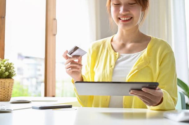 Charmante asiatische Millennial-Frau in lässigem Outfit sitzt in ihrem minimalen Arbeitsbereich, in der Hand eine Kreditkarte und ein Tablet. - Foto, Bild
