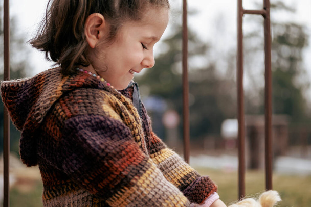 Όμορφο κοριτσάκι σε ένα καφέ πουλόβερ παίζει με την κούκλα της στον κήπο σε μια ημέρα του φθινοπώρου. Επιλεκτική εστίαση κορίτσι. ειρήνη και ευτυχία - Φωτογραφία, εικόνα
