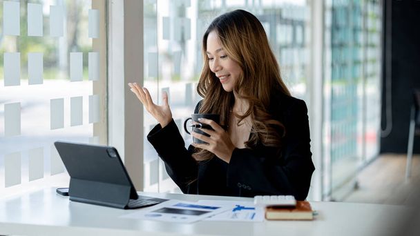 Beautiful Asian businesswoman sitting in her private office, she is talking to her partner via video call on her tablet, she is a female executive of a startup company. Concept of financial management - 写真・画像
