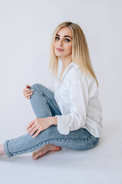 Una chica con camisa y jeans se sienta de lado sobre un fondo blanco. Fotografía de piel sin retoque. - Foto, Imagen