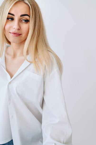 Portrait of a blonde woman who looks down to the side in a white shirt on a white background. Copy space in the right corner. - Foto, afbeelding