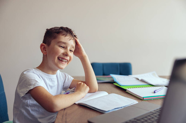 Симпатичный веселый маленький счастливый школьник с брекетами, имеющие онлайн видео урок за ноутбуком. - Фото, изображение
