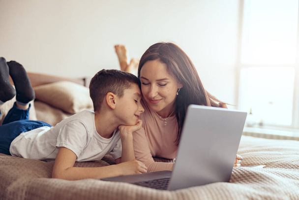 Happy Caucasian smiled mother and cute son cuddling while lying with laptop on bed. - Foto, Bild
