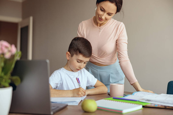 Caucasian little teen boy writing homework in copybook and mother standing by. Checking. - Photo, image
