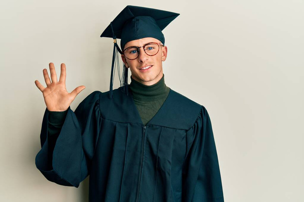 Young caucasian man wearing graduation cap and ceremony robe showing and pointing up with fingers number five while smiling confident and happy.  - 写真・画像