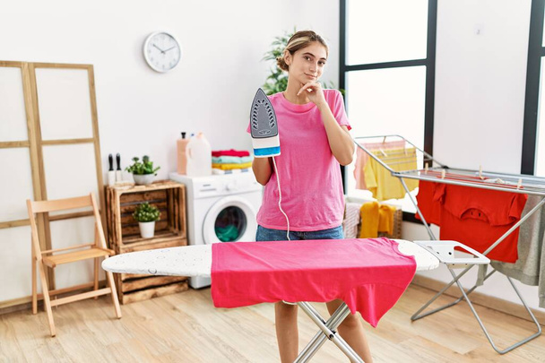 Young blonde woman ironing clothes at home serious face thinking about question with hand on chin, thoughtful about confusing idea  - Fotoğraf, Görsel