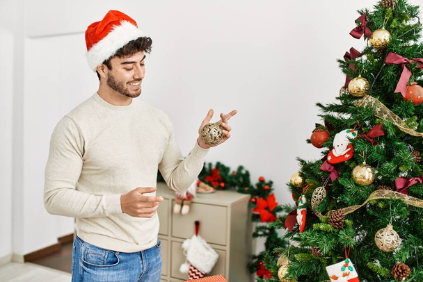 Νεαρός Ισπανός χαμογελά χαρούμενος στολίζοντας το χριστουγεννιάτικο δέντρο στο σπίτι. - Φωτογραφία, εικόνα
