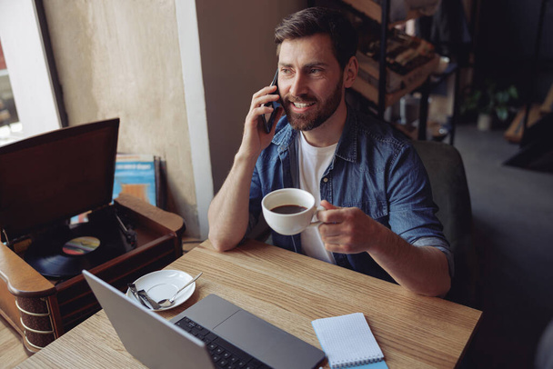 Çekici beyaz adam cep telefonuyla konuşuyor ve kafede kahve içiyor. İş sohbeti. - Fotoğraf, Görsel