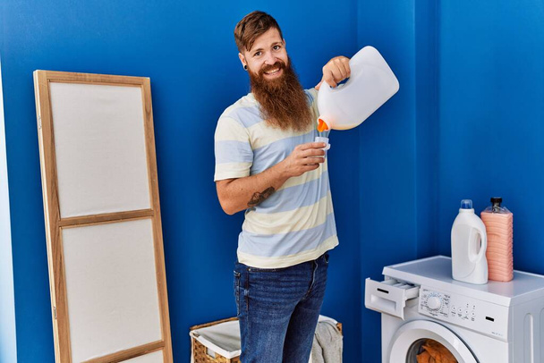 Nuori punapää mies hymyilee luottavainen kaatamalla pesuainetta pesutupa - Valokuva, kuva