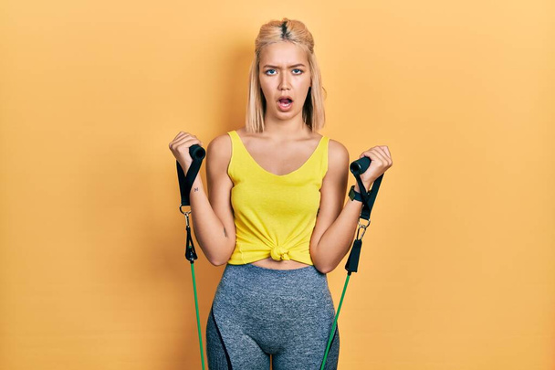 Schöne blonde Frau trainiert Armwiderstand mit elastischen Armbändern im Schockgesicht, sieht skeptisch und sarkastisch aus, überrascht mit offenem Mund  - Foto, Bild