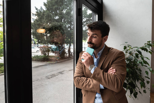 Young business man in casual suit standing near window in the restaurant holding his smartphone waiting to waiter pack the lunch to go in the box. Freelancer expat waiting take away coffee - Foto, afbeelding