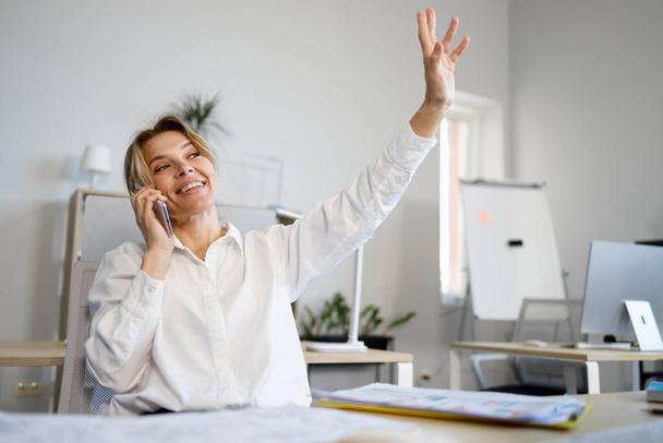 Donna d'affari positiva in camicia bianca che parla sul cellulare e agita la mano - Foto, immagini