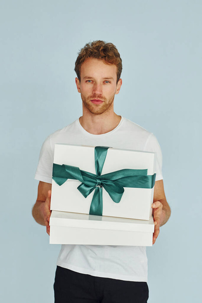 Young man in white shirt standing against wall and holds gift box. - Φωτογραφία, εικόνα