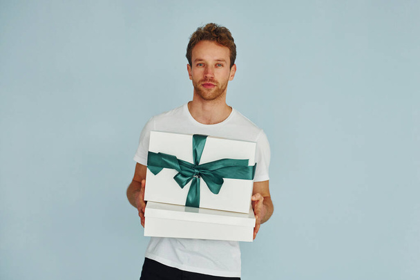 Young man in white shirt standing against wall and holds gift box. - Valokuva, kuva