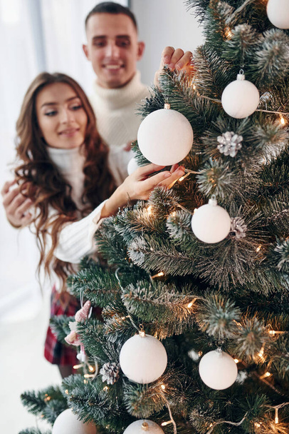 Standing near fir. Young romantic couple celebrates New year together indoors. - Φωτογραφία, εικόνα