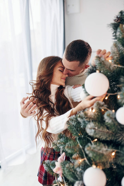 Standing near fir. Young romantic couple celebrates New year together indoors. - 写真・画像