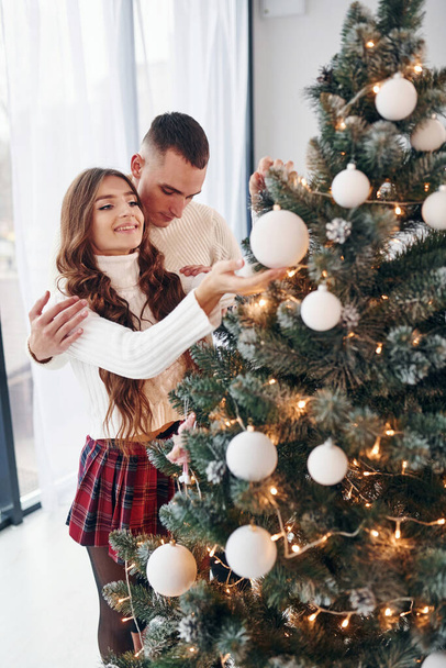 Standing near fir. Young romantic couple celebrates New year together indoors. - Φωτογραφία, εικόνα
